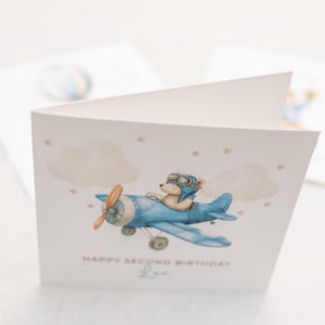 May include: A folded greeting card with a watercolor illustration of a teddy bear in a blue airplane. The card reads "HAPPY SECOND BIRTHDAY" with a name below. The background includes clouds and stars.