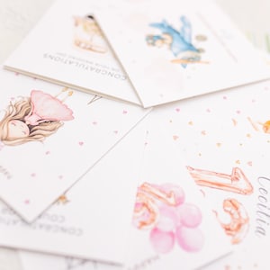 May include: A collection of greeting cards with watercolor illustrations. The cards feature images of a girl in a pink dress, a blue car, and balloons. Text includes "Happy first birthday", "Congratulations", and a name. Pink hearts are scattered on the cards.