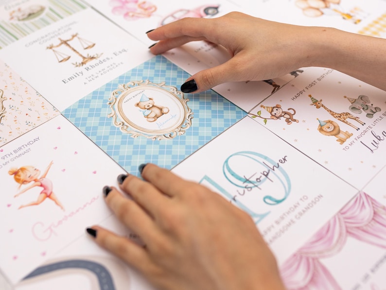 May include: A collection of greeting cards with various illustrations. One card features a teddy bear with a party hat, another has a gymnast, and others have animals. The cards are arranged on a white surface.