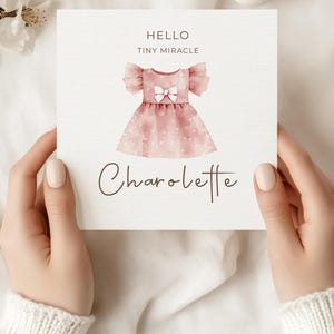 May include: A greeting card featuring a watercolor illustration of a pink dress with a bow, and the name "Charolette". The card also has the text "Hello Tiny Miracle" at the top. The card is held by a person.