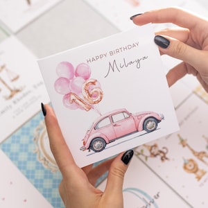 May include: A birthday card featuring a pink vintage car, pink balloons, and the number 16. The card reads "Happy Birthday Milaya" in elegant script. Other cards are visible in the background.