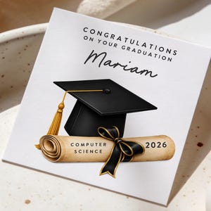 May include: A graduation card with the text "CONGRATULATIONS ON YOUR GRADUATION" and the name "Mariam". The card features a black graduation cap with a gold tassel and a scroll that reads "COMPUTER SCIENCE 2026".