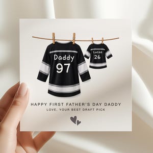 May include: A Father's Day card featuring two black and white hockey jerseys hanging on a clothesline. The larger jersey says "Daddy 97", and the smaller one says "Lucas 26". The card includes the text "HAPPY FIRST FATHER'S DAY DADDY LOVE, YOUR BEST DRAFT PICK".