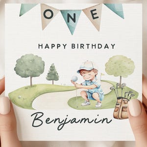 May include: A watercolor birthday card featuring a young boy playing golf. The card says "ONE" at the top, "HAPPY BIRTHDAY" in the middle, and "Benjamin" at the bottom. The boy is wearing a blue outfit and a cap.