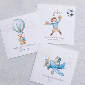 May include: Three birthday cards with watercolor illustrations. One card features a teddy bear in a hot air balloon, another shows a soccer goalie, and the third depicts a teddy bear in a biplane. Each card has a birthday message and a name.