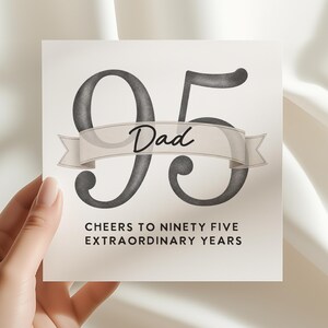 May include: A square greeting card featuring the large dark gray numbers 95 with a beige ribbon banner across the center containing the word Dad in black script. Below, the text reads CHEERS TO NINETY FIVE EXTRAORDINARY YEARS in black.