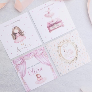 May include: Four greeting cards with illustrations of ballerinas and ballet-themed elements. The cards feature text such as "Happy First Birthday" and names like "Amelie" and "Olivia". The color palette is soft pink and white.