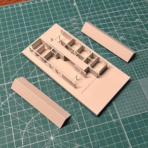 May include: A light beige architectural model with interior walls and three detached wall sections. The model is on a green cutting mat with grid lines and text. The text on the mat reads "TAMIYA CUTTING MAT".