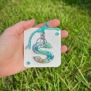 May include: A teal and sand-colored resin letter "S" keychain. The letter is filled with dried flowers and sand, attached to a silver keyring. The keychain is tied to a white card with teal string and star-shaped embellishments, set against a green grass background.