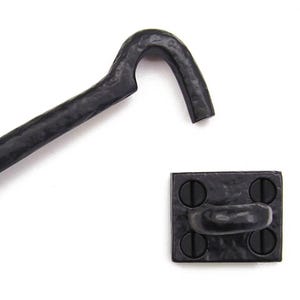 Pode incluir: Conjunto de trinco de gancho e olho de ferro preto. O gancho é curvo e preso a uma base retangular com quatro parafusos. O olho está em uma base correspondente. Ferragens rústicas para portas ou portões.