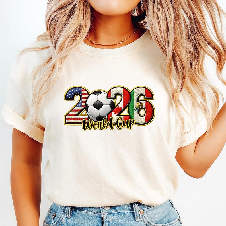 May include: Cream-colored t-shirt featuring a graphic design for the 2026 World Cup. The design includes the number 2026 with the flags of the USA, Canada, and Mexico incorporated. A soccer ball is in the center, with the words "World Cup" below.