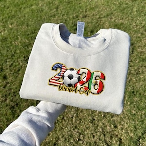 May include: Cream-colored sweatshirt with a graphic design for the 2026 World Cup. The design includes the numbers 2026, a soccer ball, and the flags of the USA, Canada, and Mexico. The words "World Cup" are also included.