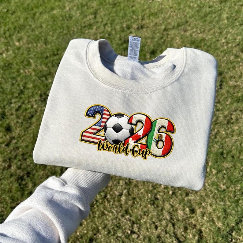 May include: Cream-colored sweatshirt with a graphic design for the 2026 World Cup. The design includes the numbers 2026, a soccer ball, and the flags of the USA, Canada, and Mexico. The words "World Cup" are also included.