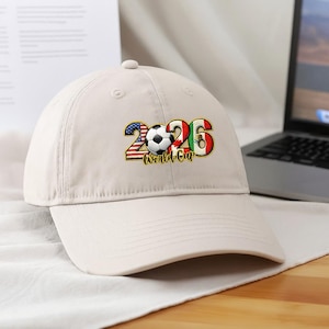 May include: A beige baseball cap featuring the text "2026 World Cup." The numbers are designed with the flags of the United States, Canada, and Mexico, with a soccer ball in the center.
