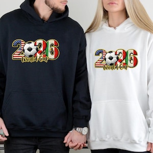 May include: Navy blue and white hooded sweatshirts with a graphic design for the 2026 World Cup. The design includes the number 2026, a soccer ball, and flags of the United States, Mexico, and Italy. The words "World Cup" are also included.