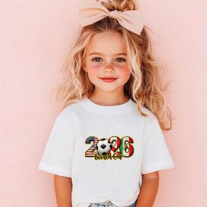 May include: A young girl wearing a white t-shirt with a soccer ball graphic. The graphic includes the text "2026 World Cup" with the numbers incorporating the American and Mexican flags. A large bow sits atop her head.