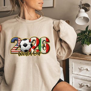 May include: Beige sweatshirt featuring a soccer ball and the text "2026 World Cup." The numbers 2 and 6 are designed with the colors of the American and Mexican flags, respectively. The soccer ball is centered between the numbers.