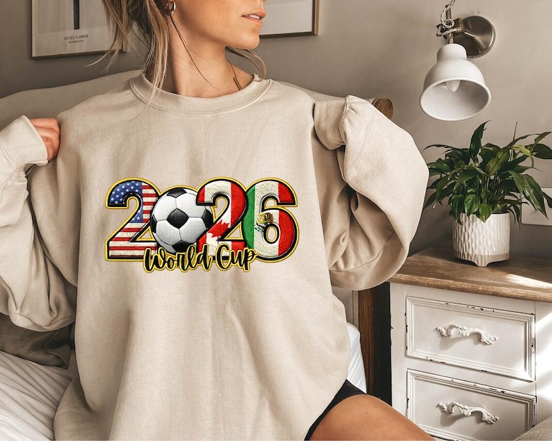 May include: Beige sweatshirt featuring a soccer ball and the text "2026 World Cup." The numbers 2 and 6 are designed with the colors of the American and Mexican flags, respectively. The soccer ball is centered between the numbers.