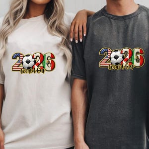 May include: Two t-shirts, one cream and one charcoal, featuring a graphic design for the 2026 World Cup. The design includes the number 2026 with the American and Mexican flags, a soccer ball, and the words "World Cup".