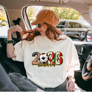 May include: Cream-colored t-shirt with a graphic design for the 2026 World Cup. The design includes the number 2026 with the American and Mexican flags, a soccer ball, and the words "World Cup".