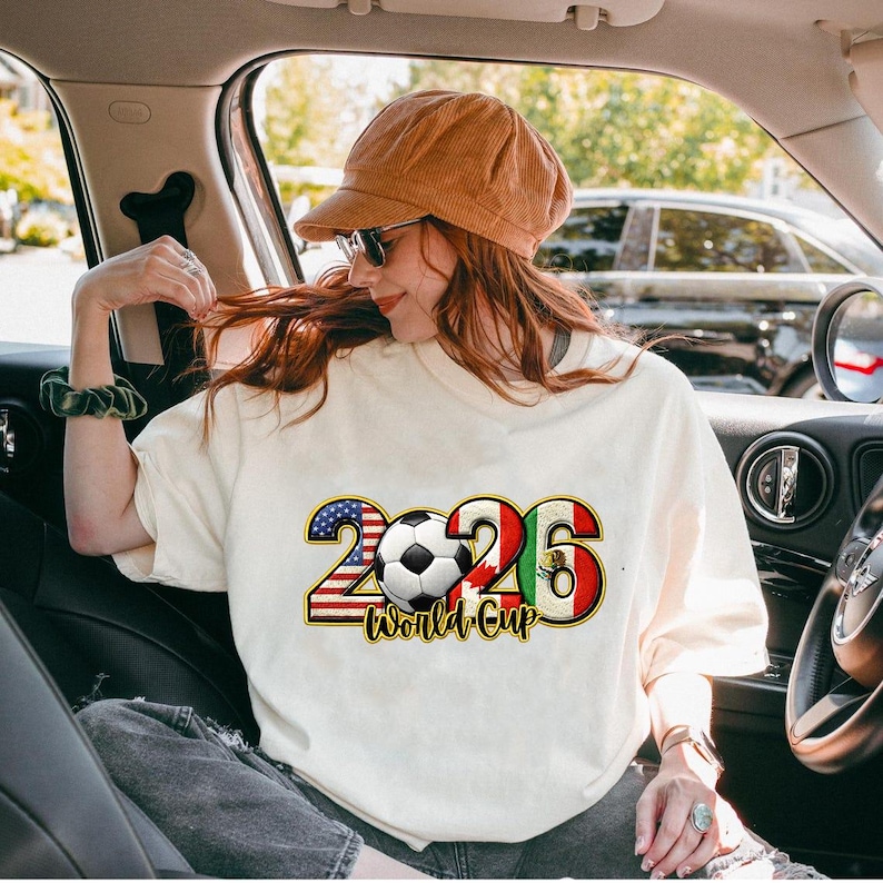 May include: Cream-colored t-shirt with a graphic design for the 2026 World Cup. The design includes the number 2026 with the American and Mexican flags, a soccer ball, and the words "World Cup".