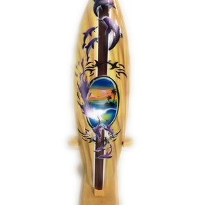 May include: A yellow wooden surfboard with a purple and blue dolphin design. The surfboard has a sunset scene with a palm tree in a circle on the front. The surfboard is on a wooden stand.