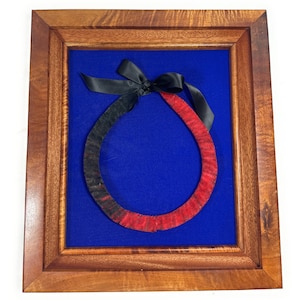 May include: A framed red and black feather lei with a black ribbon bow on a blue background.