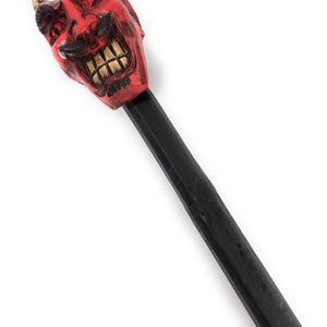 May include: A walking stick featuring a carved red devil head handle with beige horns and bared teeth. The devil face has black accents around the eyes and mouth. The shaft of the walking stick is solid black and extends downward from the head.