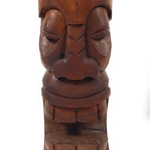 May include: A carved wooden tiki statue with a brown finish. The tiki has a large, expressive face with closed eyes and a wide mouth.