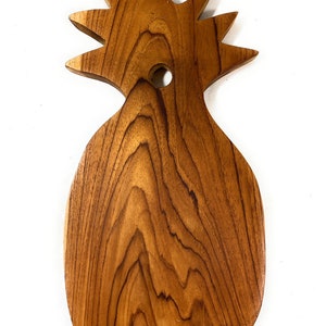 May include: A wooden pineapple-shaped cutting board with a natural wood finish. The board has a hole in the top for hanging.