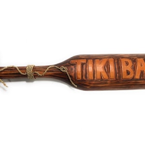 May include: A wooden Tiki Bar sign in a paddle shape. The sign is made of dark brown wood with the words "TIKI BAR" carved into it. A rope is wrapped around the handle and the paddle. The sign is designed to be hung.