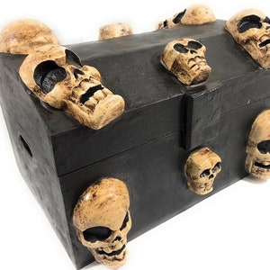 May include: A black wooden treasure chest with a hinged lid and a metal clasp. The chest is decorated with multiple skull-shaped carvings, each with a different expression.