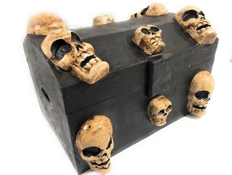 May include: A black wooden treasure chest with a hinged lid and a metal clasp. The chest is decorated with multiple skull-shaped carvings, each with a different expression.