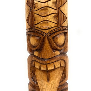 May include: A carved wooden tiki statue with a fierce expression, featuring a wide mouth with a protruding tongue and detailed eyes. The statue is brown and has a textured surface.