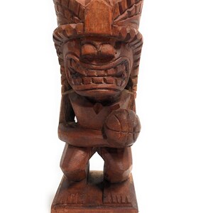 May include: A wooden tiki statue holding a basketball. The statue is carved with a fierce expression and intricate details. The basketball is also carved from wood.