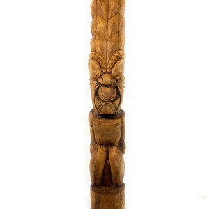 May include: A carved wooden tiki statue with a detailed design. The statue is brown and features a face with a wide mouth and a tall, textured head. It stands on a round base and has a vertical, elongated shape.