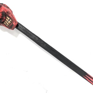 May include: A red devil head with horns and a toothy grin is attached to a black wooden walking stick with red and gold flames painted on the end.