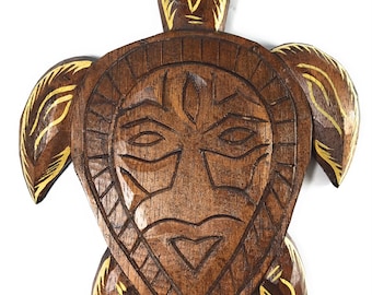 May include: A wooden turtle carving with a detailed face design. The turtle is brown with gold accents on the flippers and head. The face has carved eyes, a nose, and a mouth. The overall design is reminiscent of tribal art.