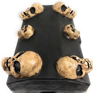 May include: A black wooden box with six skull-shaped accents. The box has a hinged lid and a small keyhole.