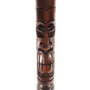 May include: A dark brown wooden tiki statue with a carved face. The face has large eyes, a wide mouth, and a carved nose.