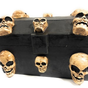 May include: A black wooden box with a hinged lid decorated with multiple sculpted human skulls. The skulls are beige and have a textured surface.