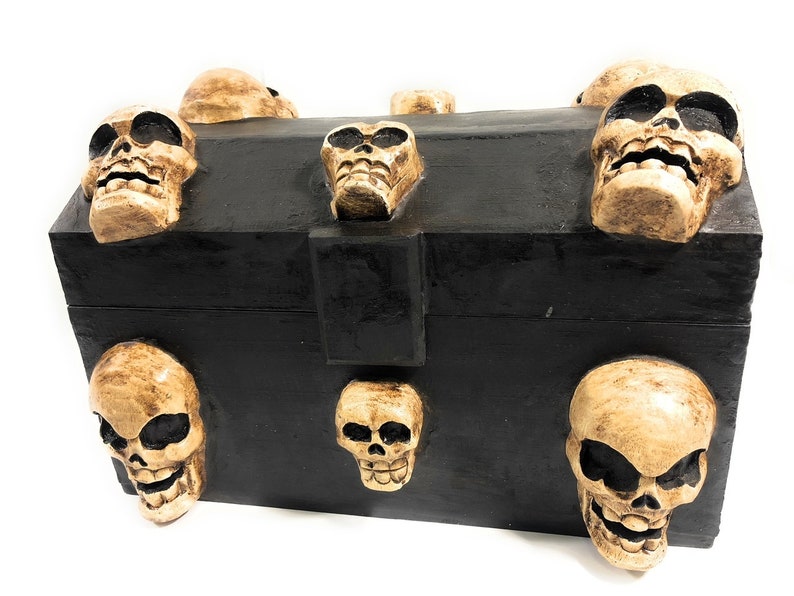 May include: A black wooden box with a hinged lid decorated with multiple sculpted human skulls. The skulls are beige and have a textured surface.