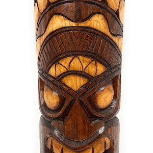 May include: A dark brown wooden tiki mask with intricate carvings. The mask has a fierce expression with large eyes, a wide mouth, and sharp teeth. The mask is decorated with geometric patterns and circles.