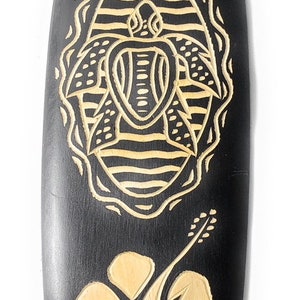 May include: Black wooden surfboard with a carved design of a turtle and hibiscus flowers. The turtle is in the center of the surfboard and the flowers are on either side of it.