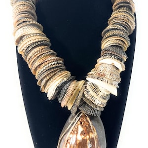 May include: A necklace featuring a series of layered, overlapping natural seashells in shades of tan, brown, and grey. The centerpiece is a large, smooth, teardrop-shaped brown and white shell encased in a thin, metallic wire frame structure.