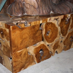 May include: A wooden coffee table made from reclaimed teak wood. The table top is made from a single piece of wood with a natural edge. The legs are made from smaller pieces of wood that have been joined together. The table has a rustic and natural look.