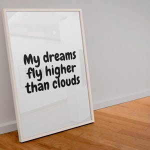 May include: A framed white print with the text "My dreams fly higher than clouds" in black cursive. The frame is leaning against a white wall on a wooden floor.