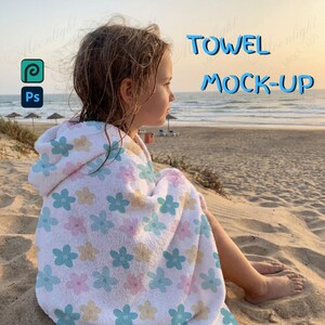 Op de afbeelding: Een kind gehuld in een witte badhanddoek met een bloemenpatroon van roze, blauwe en gele bloemen. De handdoek ligt op het strand met de tekst "TOWEL MOCK-UP" in het blauw.