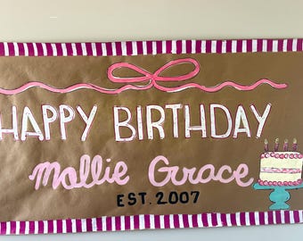 May include: A brown banner with the words "HAPPY BIRTHDAY Mollie Grace EST. 2007" in white and pink lettering. A pink bow and cake illustration are also featured. The banner is trimmed with a pink and white striped border.