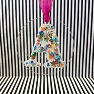 Può includere: Un ornamento circolare trasparente con la lettera "A" all'interno, decorato con gemme colorate, glitter e vernice. Un nastro rosa è attaccato per appenderlo. Lo sfondo presenta strisce bianche e nere.
